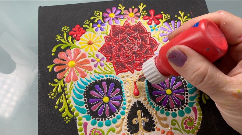 May include: A close-up of a sugar skull painting in progress. The skull is decorated with colorful flowers and dots. A hand is holding a bottle of red paint, adding details to the skull.