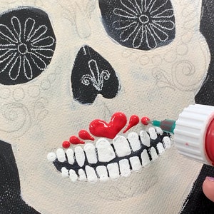 May include: A close-up of a red paint bottle being used to add details to a sugar skull painting. The skull is painted white with black details and a red heart-shaped mouth.