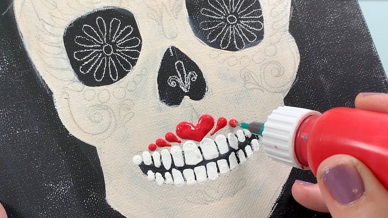 May include: A close-up of a red paint bottle being used to add details to a sugar skull painting. The skull is painted white with black details and a red heart-shaped mouth.
