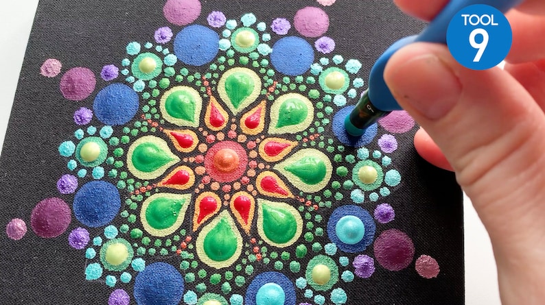 Puede incluir: Primer plano de una mano usando una herramienta de punteado azul para crear un mandala colorido en un lienzo negro. El mandala est&aacute; hecho de peque&ntilde;os puntos de varios colores, incluyendo morado, azul, verde, amarillo, naranja y rojo. La herramienta est&aacute; etiquetada como "TOOL 9".