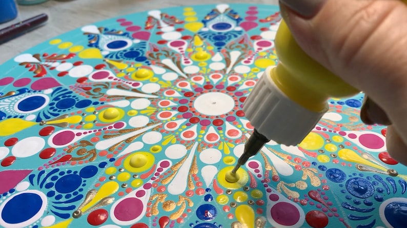 May include: A hand holding a yellow bottle of glue with a white tip, squeezing glue onto a colorful mandala painted on a round wooden surface.