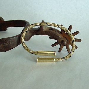 May include: A brown leather cowboy spur with a gold-colored metal wheel and a beige and black rope bracelet with two gold-colored bullet casings.