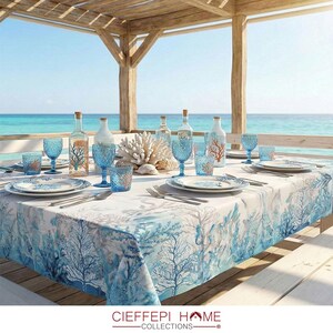 Puede incluir: Una mesa bellamente decorada con un tema costero, con un mantel blanco adornado con patrones de coral azul. La mesa está decorada con cristalería, platos y botellas azules, creando una experiencia gastronómica elegante. La escena está ambientada al aire libre con vistas al mar.