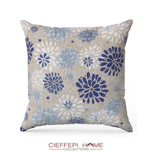 Può includere: Un cuscino decorativo con uno sfondo in lino beige e un motivo floreale blu e bianco. Il cuscino presenta un motivo ripetitivo di fiori stilizzati in tonalità di blu e bianco.