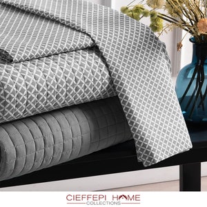 Könnte beinhalten: Ein Stapel grauer und weiß gemusterter Steppdecken mit geometrischem Design. Die Steppdecken sind gefaltet und auf einer schwarzen Oberfläche angeordnet. Der Text "CIEFEEPI HOME COLLECTIONS" ist am unteren Rand des Bildes sichtbar.