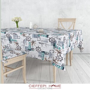 Op de afbeelding: Een wit tafelkleed met een nautisch thema met daarop afbeeldingen van vuurtorens, palmbomen, schepen en stuurwielen in tinten blauw en groen. Het tafelkleed ligt op een houten tafel met twee houten stoelen.