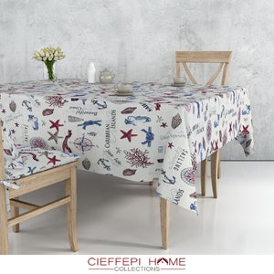 Puede incluir: Un mantel blanco con temática náutica, con ilustraciones rojas, azules y beige de anclas, estrellas de mar, conchas y brújulas. El mantel cubre una mesa y una silla de madera, creando una estética costera.