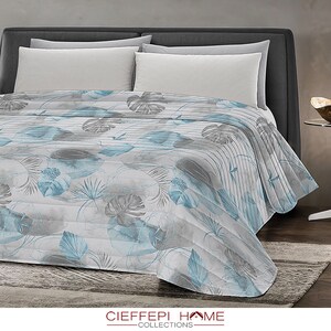 Könnte beinhalten: Eine weiße und blaue gemusterte Steppdecke mit einem dezenten Aquarell-Effekt. Die Steppdecke zeigt ein Design aus Blättern und Blumen in Blautönen und Grau. Die Steppdecke ist auf einem Bett mit weißen Kissen gefaltet.