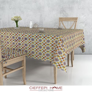 May include: A white tablecloth with a colorful geometric pattern in shades of yellow, blue, orange, and red. The tablecloth is draped over a wooden table with two wooden chairs.