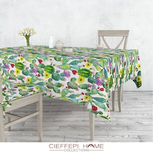 Può includere: Una tovaglia bianca con un motivo di cactus e fiori verdi. La tovaglia è su un tavolo di legno con due sedie di legno.