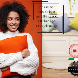 Può includere: Una donna tiene in mano un cuscino arancione brillante. Il testo sul muro recita "Tessuto morbido anallergico antiacaro traspirante". L'immagine mostra anche un divano bianco con cuscini e un tappeto.