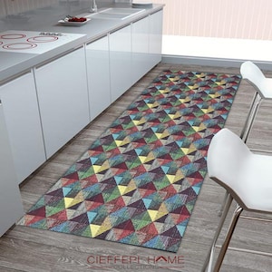 May include: A colorful geometric runner rug with a pattern of triangles in shades of red, blue, green, yellow, and brown. The rug is placed in a kitchen setting with white cabinets and a wood floor.