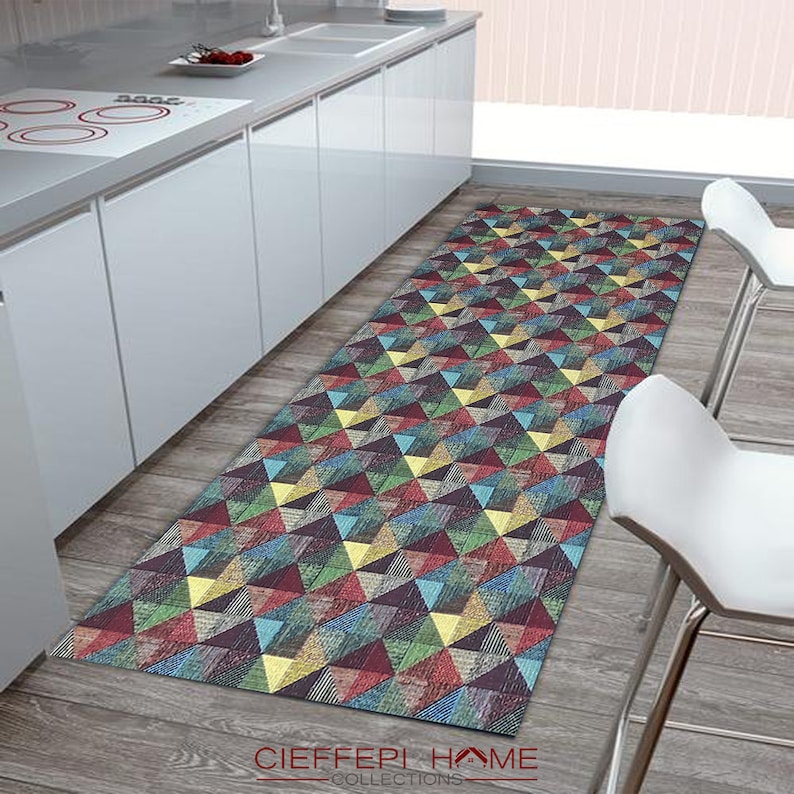 May include: A colorful geometric runner rug with a pattern of triangles in shades of red, blue, green, yellow, and brown. The rug is placed in a kitchen setting with white cabinets and a wood floor.