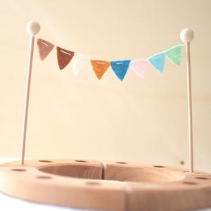 Peut inclure: Une base circulaire en bois avec de multiples trous, dotée de deux tiges verticales en bois surmontées de sphères. Une guirlande de petits fanions triangulaires en feutrine aux tons marron, blanc, orange, bleu, rose et vert clair est suspendue.