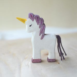 May include: A white wooden unicorn figurine with a yellow horn, purple mane, and tail. The unicorn has a simple, handcrafted design with painted details. The hooves are painted a dark pink color. The figurine is displayed on a light-colored wooden surface.
