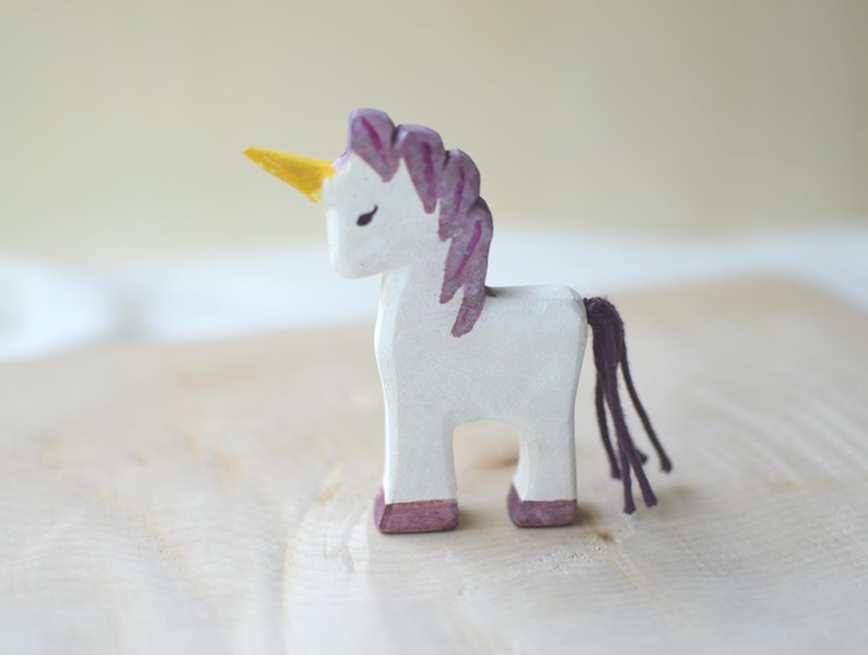 May include: A white wooden unicorn figurine with a yellow horn, purple mane, and tail. The unicorn has a simple, handcrafted design with painted details. The hooves are painted a dark pink color. The figurine is displayed on a light-colored wooden surface.