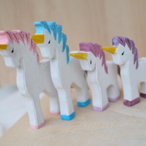 May include: Four wooden unicorn figurines with white bodies, colorful manes, and yellow horns. The manes are pink, blue, purple, and pink. The unicorns stand on a light-colored wooden surface.