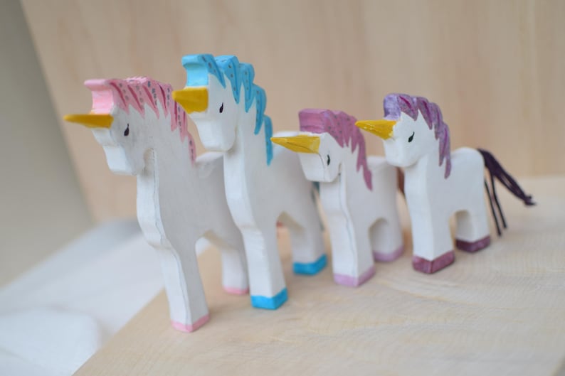 May include: Four wooden unicorn figurines with white bodies, colorful manes, and yellow horns. The manes are pink, blue, purple, and pink. The unicorns stand on a light-colored wooden surface.