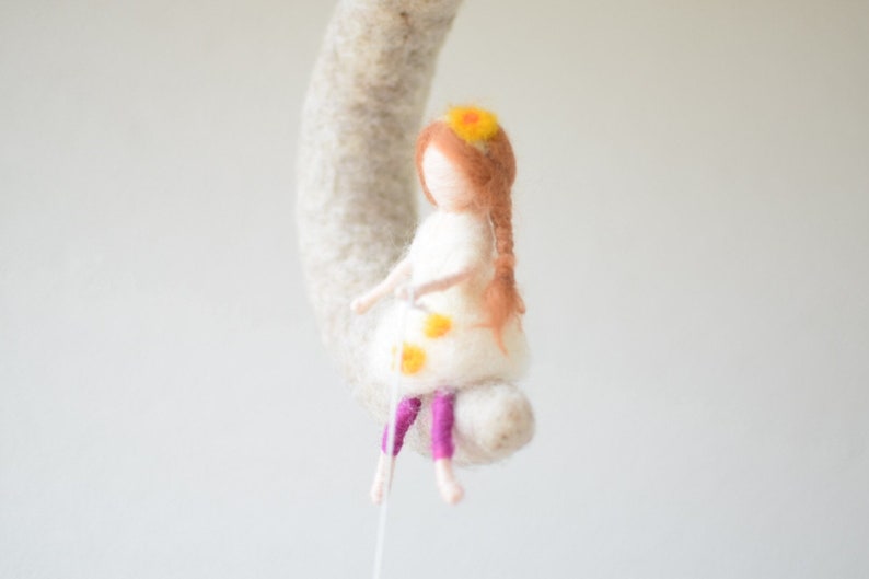 May include: A felt doll with brown hair and a white dress with yellow flowers. The doll is wearing purple pants and has a yellow flower crown.