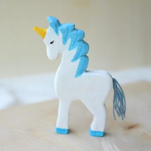 May include: A handcrafted wooden unicorn figurine. The unicorn is painted white with a blue mane and tail, a yellow horn, and blue hooves. The toy is standing on a wooden surface.