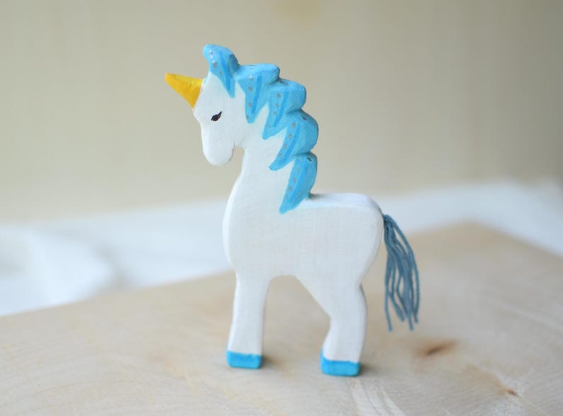 May include: A handcrafted wooden unicorn figurine. The unicorn is painted white with a blue mane and tail, a yellow horn, and blue hooves. The toy is standing on a wooden surface.