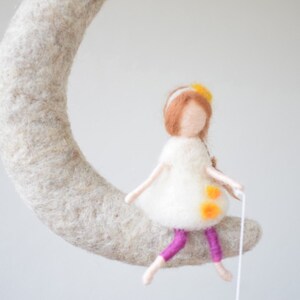 May include: A felt doll with brown hair and a white dress sits on a felt crescent moon. The doll has yellow flowers in her hair and is wearing purple leggings.