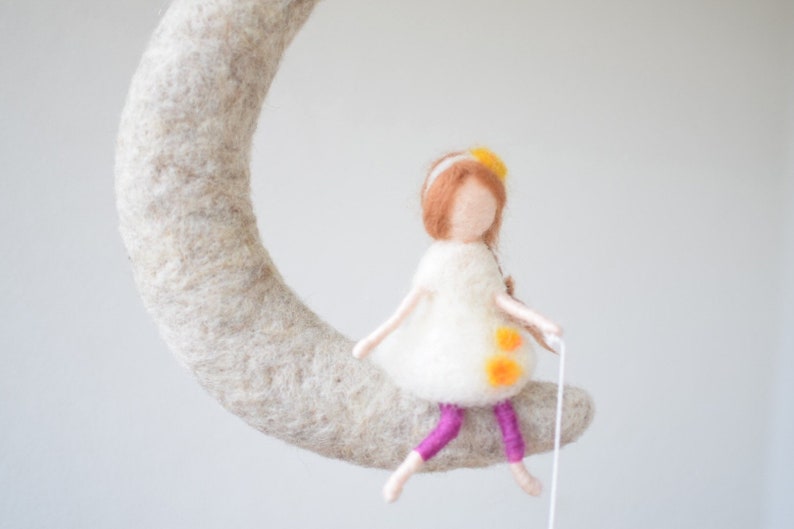 May include: A felt doll with brown hair and a white dress sits on a felt crescent moon. The doll has yellow flowers in her hair and is wearing purple leggings.