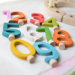 Pu&ograve; includere: Una collezione di dieci numeri in legno da zero a nove, ognuno dipinto in un colore distinto tra cui rosa, arancione, rosso, verde lime, bianco, ottanio, giallo e azzurro. Ogni numero presenta un piccolo piolo cilindrico in legno alla base.