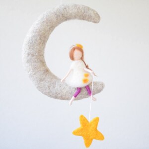 May include: A felt moon mobile with a girl doll sitting on it and holding a yellow felt star. The girl doll has brown hair, a white dress with yellow dots, and pink leggings.