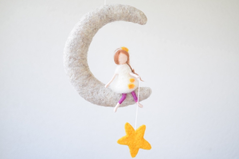 May include: A felt moon mobile with a girl doll sitting on it and holding a yellow felt star. The girl doll has brown hair, a white dress with yellow dots, and pink leggings.