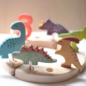 May include: A collection of colourful wooden dinosaur toys, including blue, green, and purple, sit on a light wooden base. A red number 3 is also present. The toys are designed with a simple, child-friendly aesthetic.