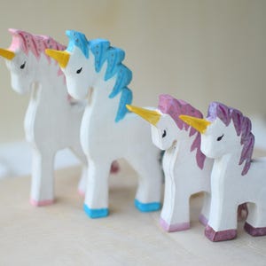 May include: Four hand-carved wooden unicorn figurines. The unicorns have white bodies, yellow horns, and colorful manes and hooves in pink, blue, and purple. They are arranged on a light-colored wooden surface.
