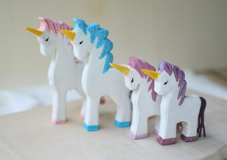 May include: Four hand-carved wooden unicorn figurines. The unicorns have white bodies, yellow horns, and colorful manes and hooves in pink, blue, and purple. They are arranged on a light-colored wooden surface.