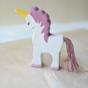 May include: A handcrafted wooden unicorn figurine. The toy is painted white with a pink mane, tail, and hooves. It has a yellow horn. The unicorn stands on a light-colored wooden surface.