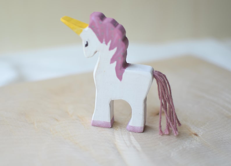 May include: A handcrafted wooden unicorn figurine. The toy is painted white with a pink mane, tail, and hooves. It has a yellow horn. The unicorn stands on a light-colored wooden surface.