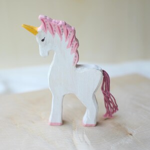 May include: A white wooden unicorn figurine with a pink mane and tail, and a yellow horn. The unicorn is standing on a light-colored wooden surface.