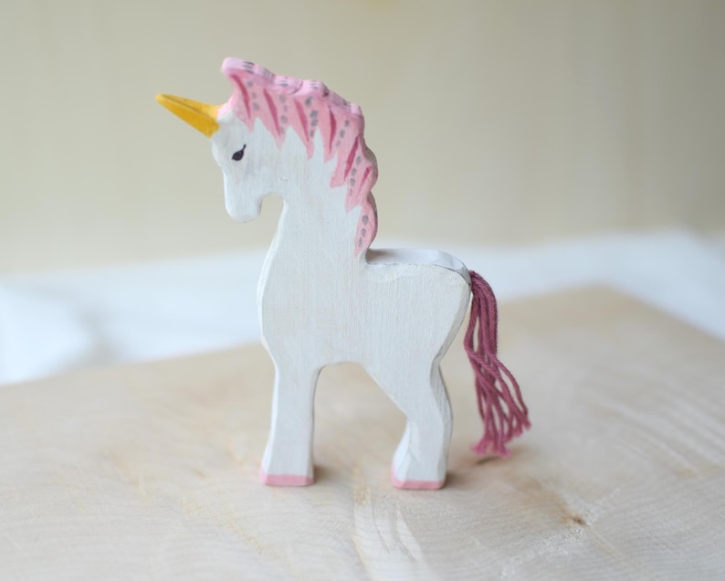 May include: A white wooden unicorn figurine with a pink mane and tail, and a yellow horn. The unicorn is standing on a light-colored wooden surface.