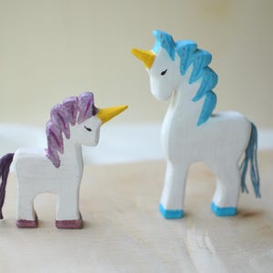 May include: Two wooden unicorn figurines. One has a purple mane and tail, the other has a blue mane and tail. Both have yellow horns and are painted white. The unicorns are standing on a light-colored wooden surface.