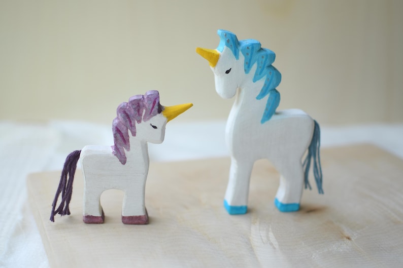 May include: Two wooden unicorn figurines. One has a purple mane and tail, the other has a blue mane and tail. Both have yellow horns and are painted white. The unicorns are standing on a light-colored wooden surface.