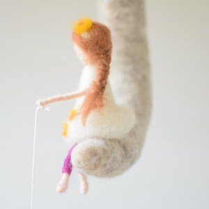 May include: A felt doll with long brown hair and a white dress. The doll is holding a string and has a yellow flower crown on its head. The doll is hanging from a white felt cloud.