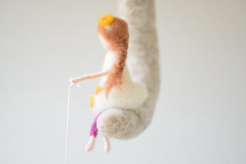 May include: A felt doll with long brown hair and a white dress. The doll is holding a string and has a yellow flower crown on its head. The doll is hanging from a white felt cloud.