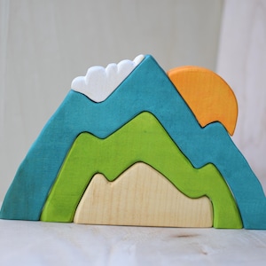 May include: A wooden mountain puzzle with a teal, green, and natural wood color scheme. The puzzle features a sun and clouds, and is designed to be stacked. The mountain is approximately 6 inches tall.
