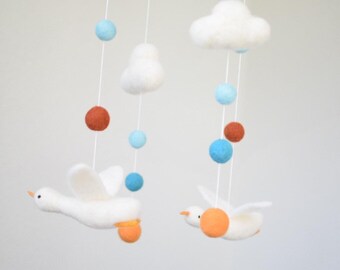 May include: A handmade baby mobile featuring two white felt geese with orange feet and beaks, suspended below felt clouds and colourful felt balls in shades of blue and rust. The nursery decor is suspended by white strings.