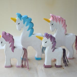 May include: Four wooden unicorn figurines with white bodies, colorful manes, and golden horns. The manes are painted in shades of blue, pink, and purple. The unicorns stand on a wooden surface.