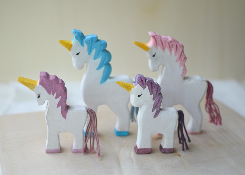 May include: Four wooden unicorn figurines with white bodies, colorful manes, and golden horns. The manes are painted in shades of blue, pink, and purple. The unicorns stand on a wooden surface.