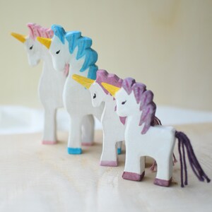 May include: Four wooden unicorn figurines with white bodies, colorful manes, and tails. The manes are pink, blue, and purple, with yellow horns and pink, blue, and purple hooves. The unicorns are standing on a light-colored wooden surface.