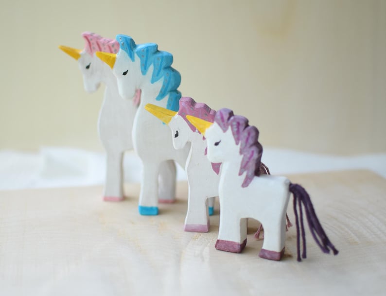 May include: Four wooden unicorn figurines with white bodies, colorful manes, and tails. The manes are pink, blue, and purple, with yellow horns and pink, blue, and purple hooves. The unicorns are standing on a light-colored wooden surface.