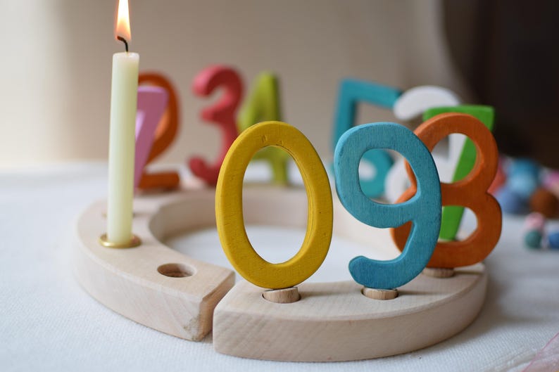 Pu&ograve; includere: Una base circolare in legno per compleanni con diversi fori per gli inserti. Una candela color crema accesa &egrave; inserita in un foro. Diversi numeri in legno colorati, tra cui uno zero giallo, un nove blu e un otto arancione, sono inseriti.