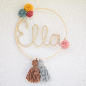 以下が含まれることがあります： ベージュの糸で「Ella」という名前が書かれた木製フープ。フープはピンク、イエロー、ブルーのカラフルなポンポンで飾られています。フープの下部から2つのタッセルがぶら下がっています。1つはブラウン、もう1つはグレーです。