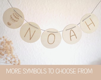 Peut inclure: Guirlande en bois avec le nom "NOAH" écrit sur des disques ronds. Un disque est orné d'un motif de montgolfière. La guirlande est suspendue à un fil fin. Le texte "MORE SYMBOLS TO CHOOSE FROM" est en bas.